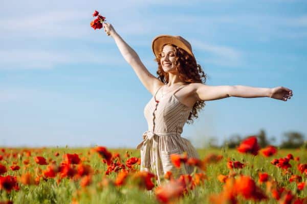 Mulher bonita em meio a um campo de flores vermelhas, usando um vestido leve e chapéu, segura algumas flores nas mãos.  Conceito de tratamentos estéticos realizados na primavera.