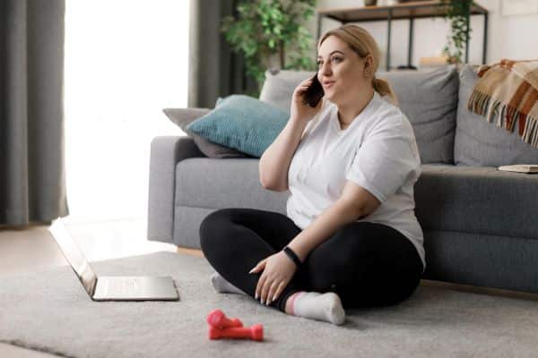 Mulher obesa, sentada no tapete cinza frente ao sofá, conversa pelo telefone celular, enquanto usa um notebook.