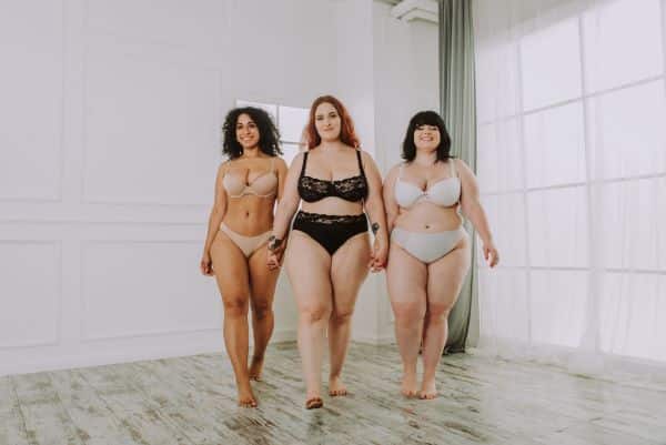 Três diferentes mulheres, com acúmulo de gordura em distintas partes do corpo, caminham por uma ampla sala para serem fotografadas. Conceito de lipedema e obesidade feminina.
