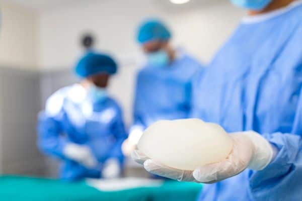 Sala de cirurgia na qual se encontram duas auxiliares e o médico cirurgião. Uma das mulheres segura uma prótese de silicone que será inserida nas mamas da paciente.