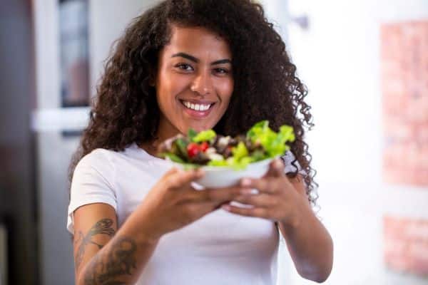 Mulher negra, jovem, com cabelos encaracolados e sorridente mostra um prato cheio de verduras e legumes. 
Alusão à alimentação saudável.