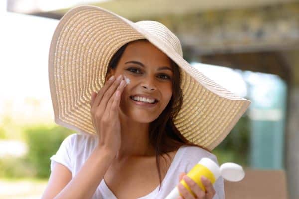 Linda mulher com cabelos escuros e longos protege o rosto com protetor solar facial e chapéu de abas largas.