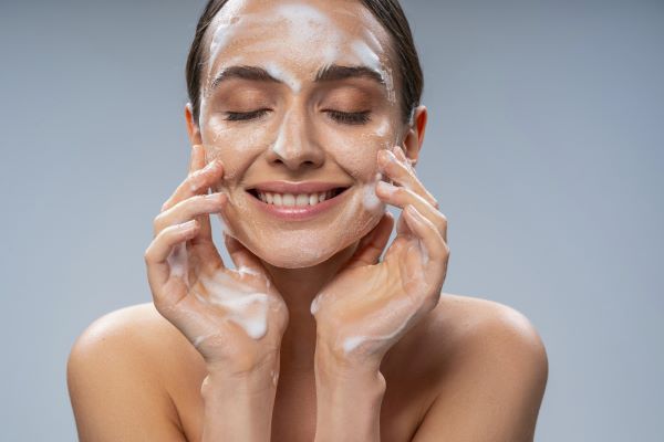 Mulher bonita, com cabelos escuros presos atrás da nuca, faz uma limpeza na pele da face com sabonete hidratante.