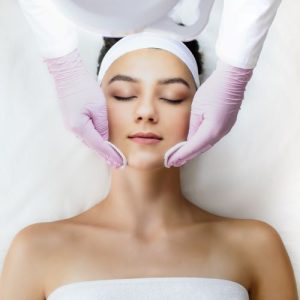 Paciente jovem e bonita realizando peeling facial com profissional que usa luvas rosas e passa algodões por sua face.