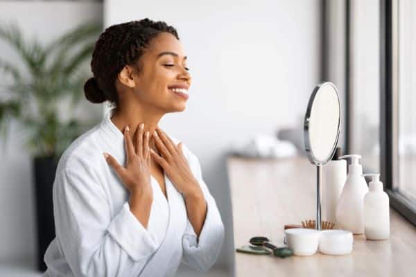 Linda mulher negra, sorridente, vestindo um robe branco, aprecia-se no espelho de mesa redondo frente a ela. Sobre a bancada, cremes e produtos de beleza.