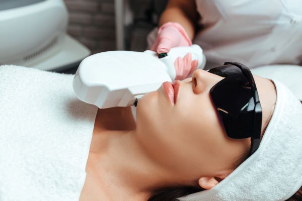 Close de uma mulher jovem realizando tratamento facial com luz pulsada. Ela usa óculos escuros de proteção ocular, está com uma toalha branca cobrindo o peito e uma faixa branca prendendo os cabelos.