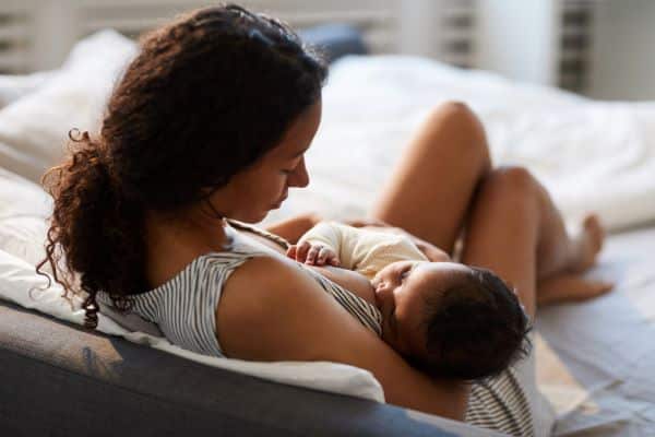 Jovem mamãe recostada no sofá, com cabelos encaracolados e presos em rabo de cavalo, está amamentando seu bebê.