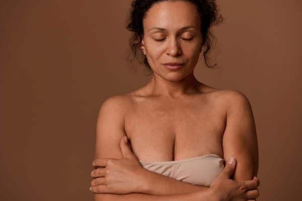 Close-up de uma mulher serena posando com os olhos fechados e cruzando os braços frente aos seios que estão cobertos por um sutiã. Imagem destaca o processo de envelhecimento de mamas.