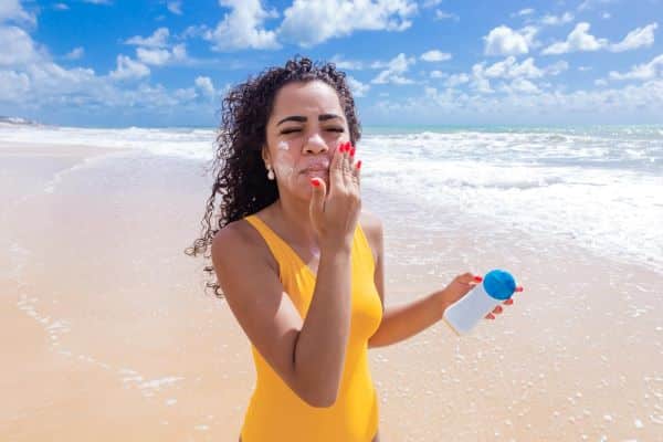 Bela mulher negra aproveita o sol na praia e protege-se dos raios nocivos passando protetor solar no rosto.