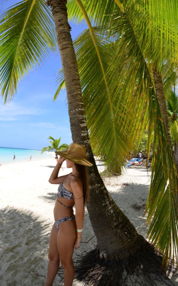 Mulher jovem, vestindo biquíni e usando um chapéu, protege sua pele dos efeitos nocivos dos raio UV aproveitando uma sombra embaixo de coqueiros na praia.