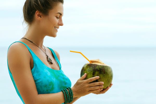 Mulher jovem, vestindo uma regata azul e com os cabelos presos, sorri segurando um coco frente à praia.