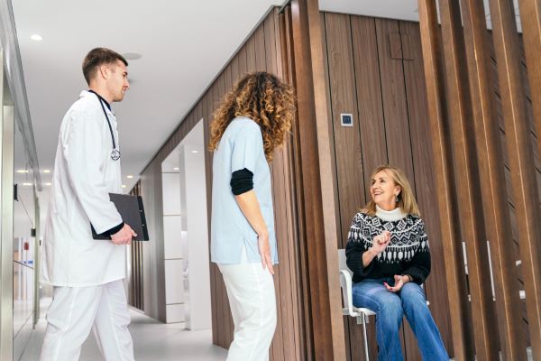 Mulher, sentada em uma poltrona da sala de espera do hospital, conversa com técnica de enfermagem e médico antes de dirigir-se à sua cirurgia plástica.