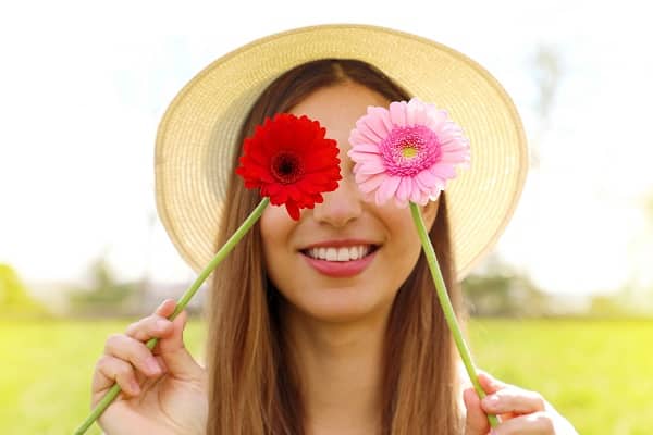 Jovem loira, com cabelos compridos, passeia pelo campo usando um chapéu e cobrindo os olhos com 2 gérberas (vermelha e rosa). 

Conceito de cirurgias plásticas na primavera.

