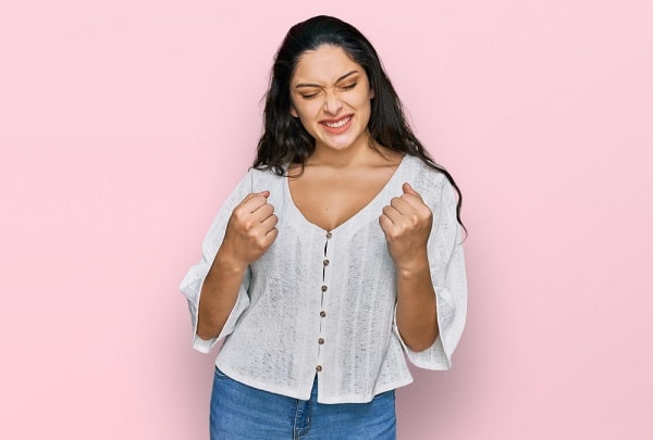 Garota com cabelos cacheados e escuros, feliz, faz o gesto de vitória com os punhos cerrados e os braços erguidos, após ter feito mamoplastia redutora.