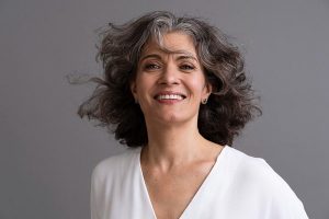 Linda mulher madura, com cabelos encaracolados e grisalhos, vestindo camisa branca, sorri para a câmera.