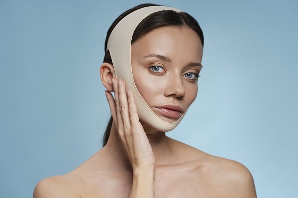 Bela mulher de olhos claros, usando faixa cirúrgica no rosto no pós-operatório de lifting facial.