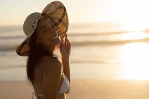 Linda jovem com cabelos longos e escuros, usando um biquini branco e um chapéu passeia pela beira da praia no pôr do sol.