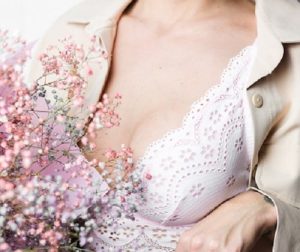 Mulher segurando um pequeno buquê de flores rosas miúdas, vestindo uma camisa aberta que deixa os seios semi amostra com um sutiã branco rendado.