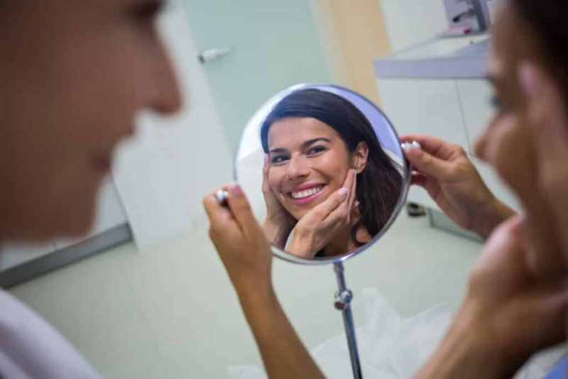 Paciente refletida em um espelho, sorri enquanto médica verifica o resultado do procedimento.