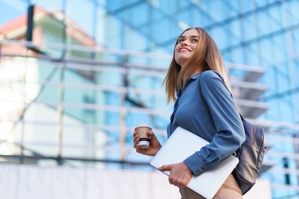 Um dos mitos do pós-operatório é a volta ao trabalho em 7 dias. Isso depende do procedimento.
Na imagem, uma linda jovem, vestindo camisa azul , calça bege e mochila de couro, segura um copo de café e um notebook.