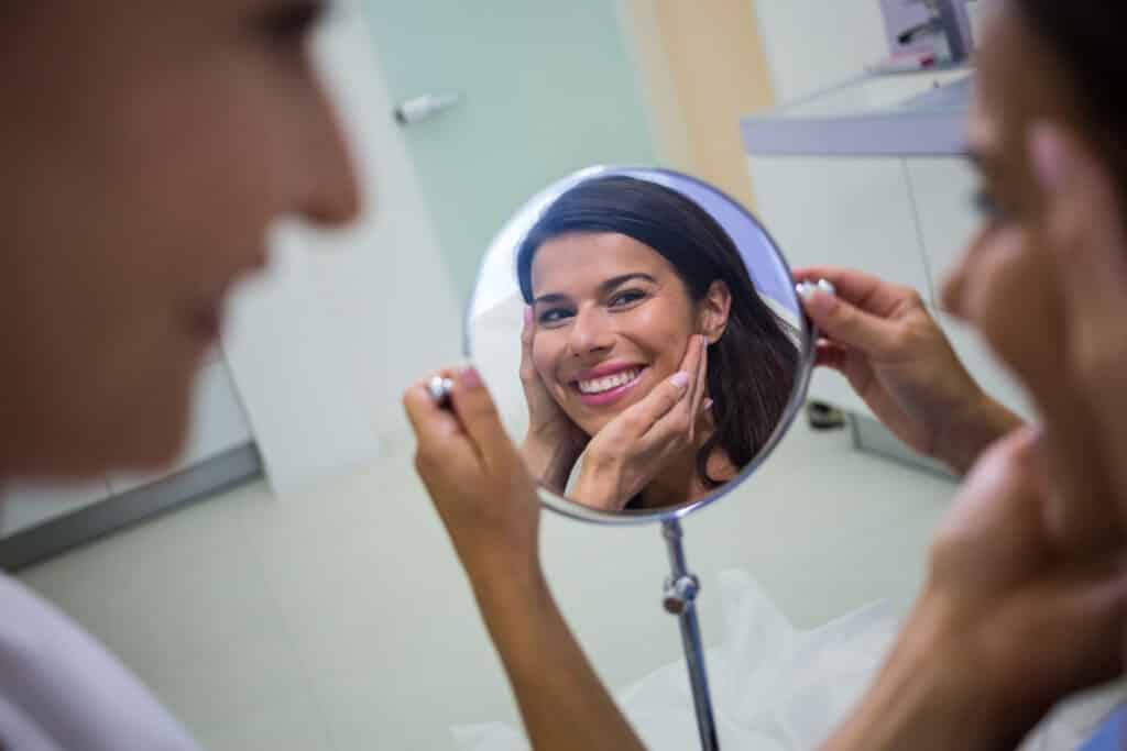 Paciente refletida em um espelho, sorri enquanto médica verifica o resultado do procedimento.