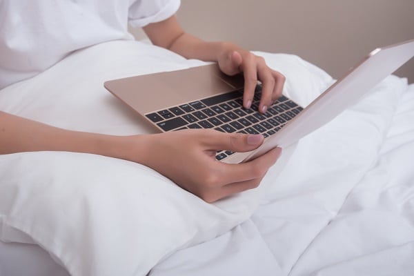 Paciente acamado mexendo no notebook. A roupa de cama é toda branca, assim como a camiseta do mesmo.